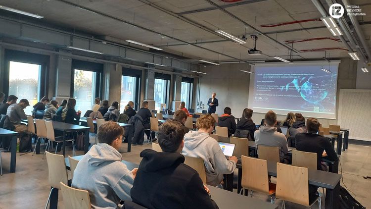 Zdjęcie z sali podczas prelekcji. Na pierwszym planie studenci, dalej widać ekran z prezentacją oraz gość pan  Ireneusz Kozber, Dyrektor Logistyki w Millano Group