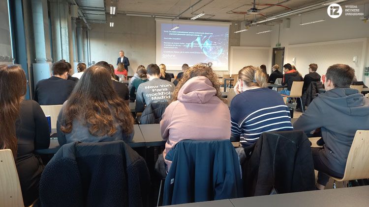 Zdjęcie z sali podczas prelekcji. Na pierwszym planie studenci, dalej widać ekran z prezentacją oraz gość pan  Ireneusz Kozber, Dyrektor Logistyki w Millano Group